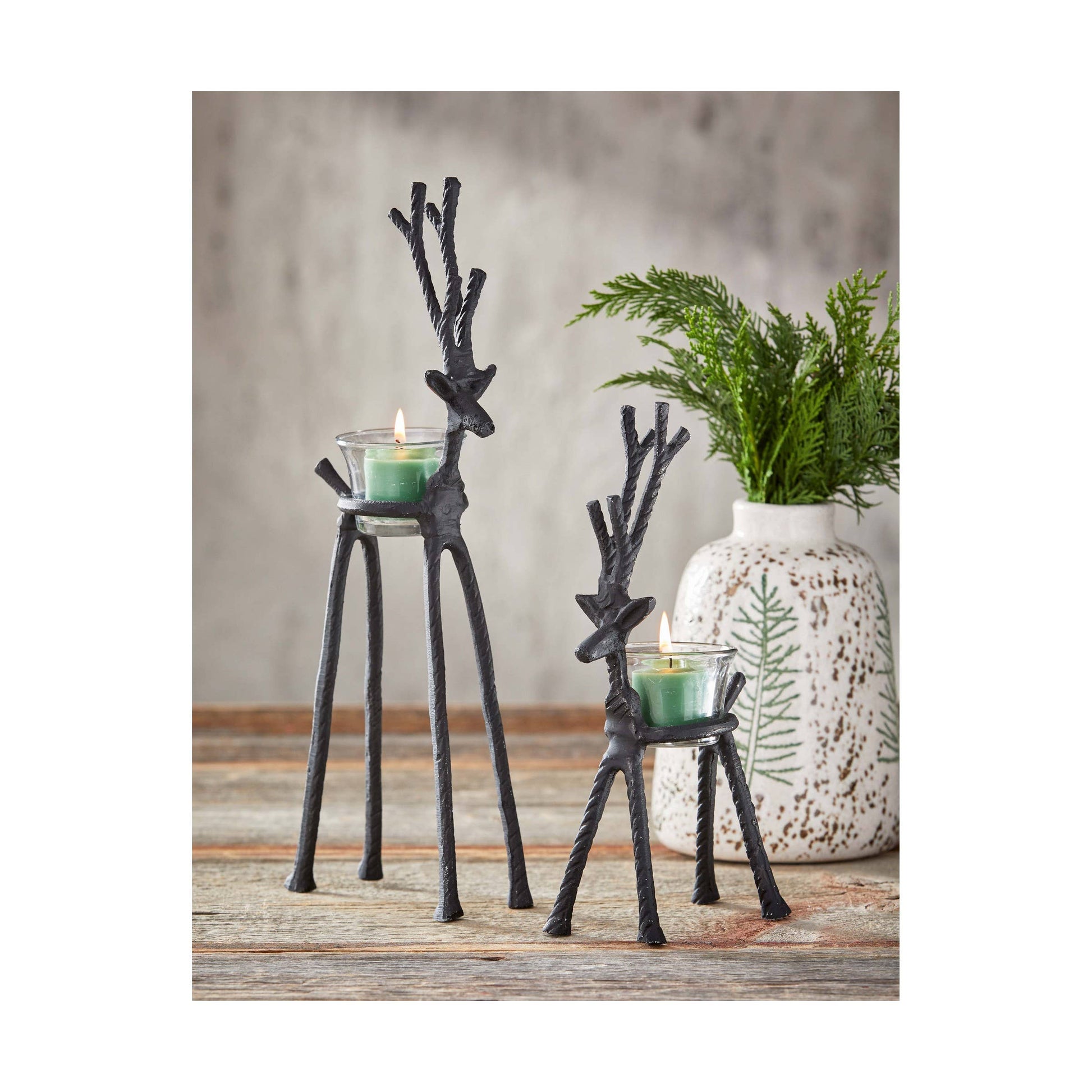 Two black metal deer candle holders with green candles on a wooden surface.