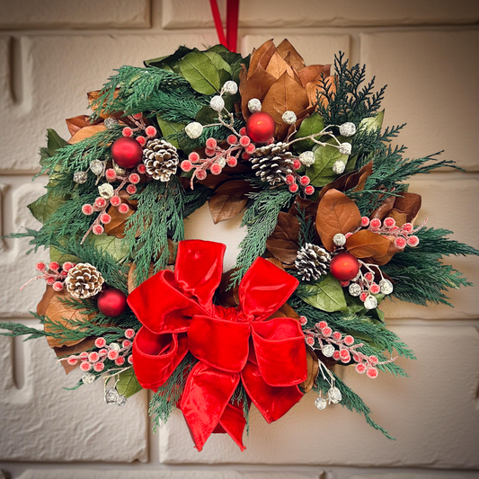Decorative Christmas wreath with red bow, pine cones, and berries on a white wall.