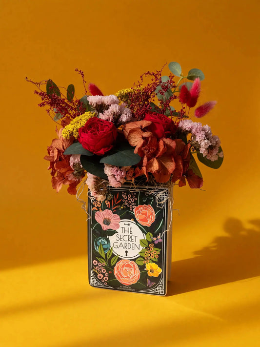 A floral arrangement in a decorative book-shaped vase with the title 'The Secret Garden' printed on the front, set against a solid warm yellow background.