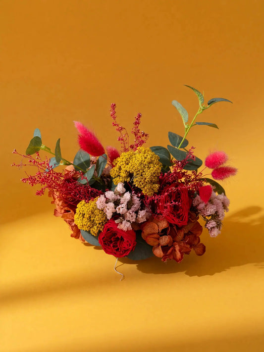 A vibrant artificial floral arrangement in a dark green bowl, featuring red roses, yellow clusters, pink fluffy stems, and green leaves, set against a solid mustard yellow background.