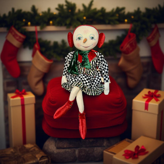 Joe Spencer art doll in a checkered outfit sitting on a red cushion with Christmas stockings and presents in the background.