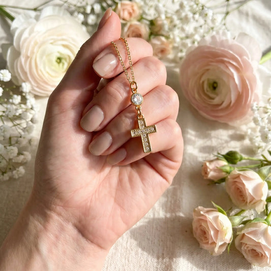 Hand holding a gold necklace with a cross pendant against a floral background