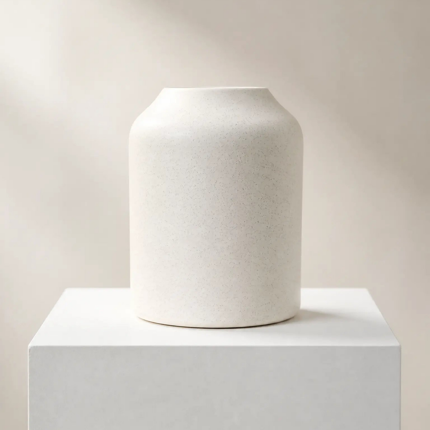 A matte off-white ceramic vase with a flared rim, displayed on a matching white pedestal against a warm beige background with soft diagonal shadows.