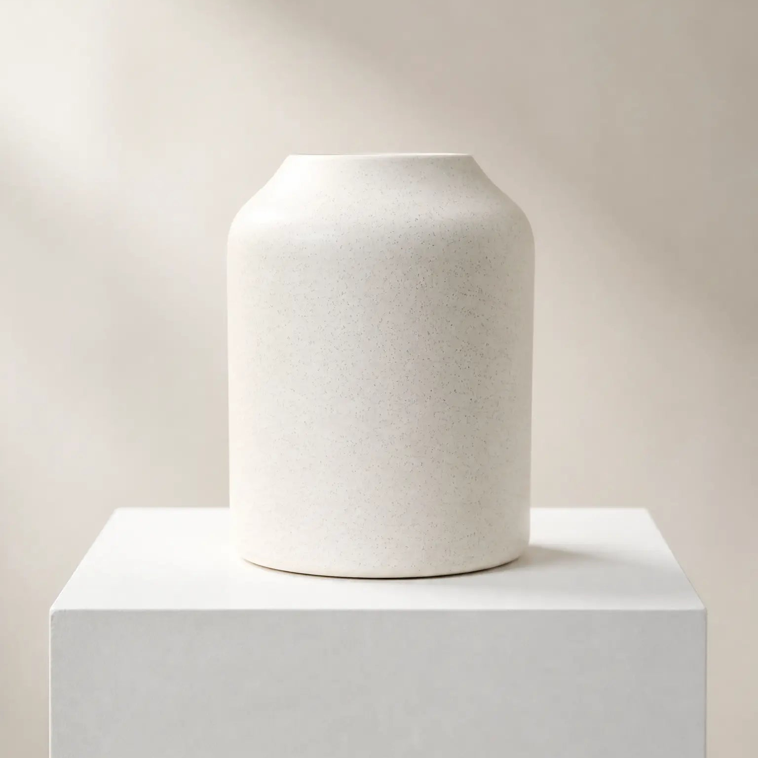 A matte off-white ceramic vase with a flared rim, displayed on a matching white pedestal against a warm beige background with soft diagonal shadows.