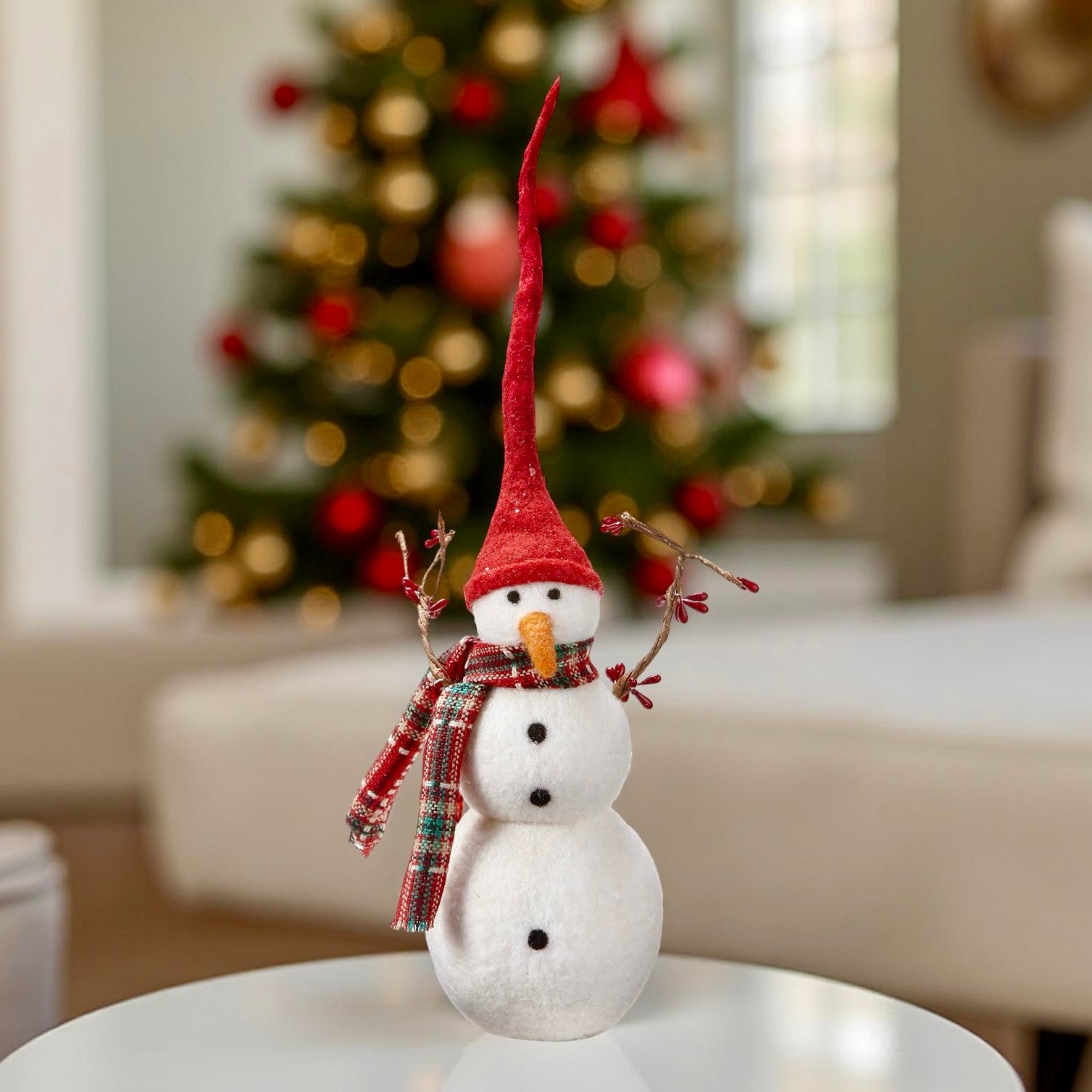 Decorative snowman with a red hat and plaid scarf on a table with a Christmas tree in the background.